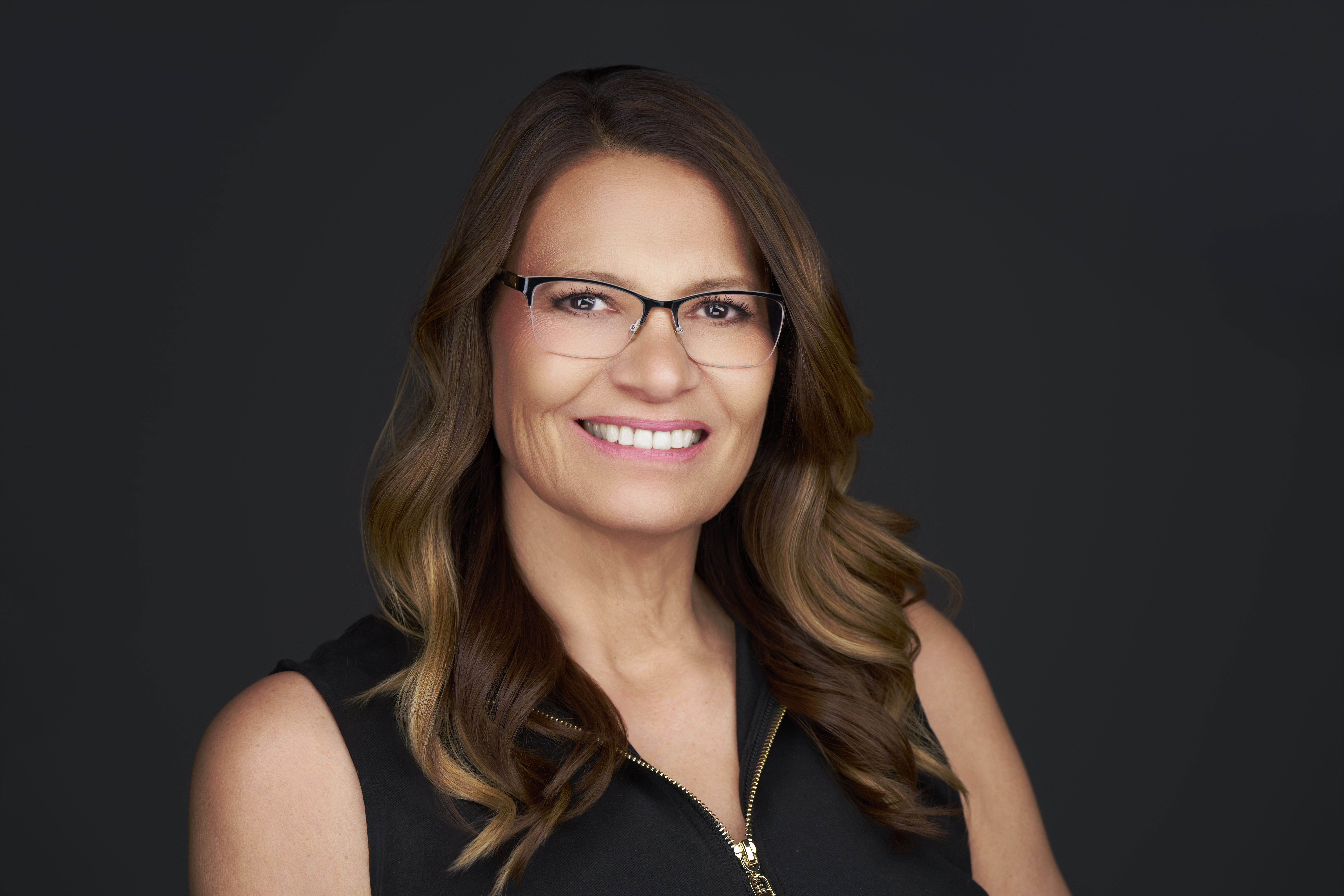Woman with long brown hair, wearing glasses, a dark pinstripe blazer, light blouse, and necklace, smiling in front of a mottled grey background.